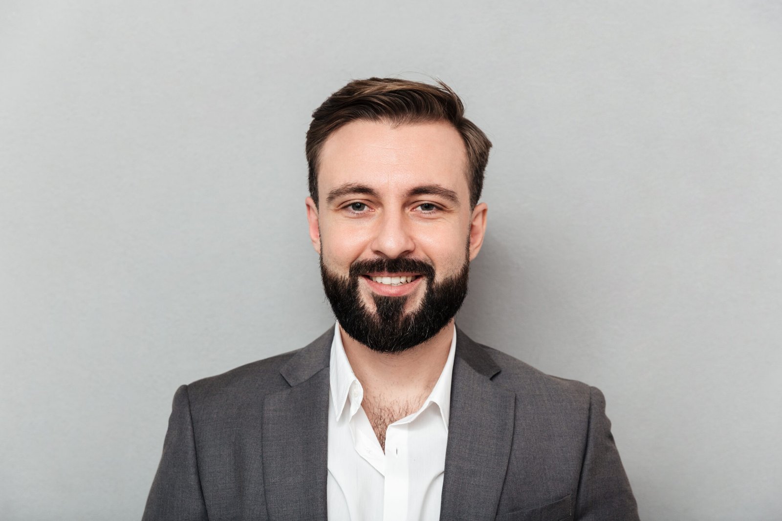Close up portrait of young bearded man in white shirt and jacket posing on camera with broad smile isolated over gray background